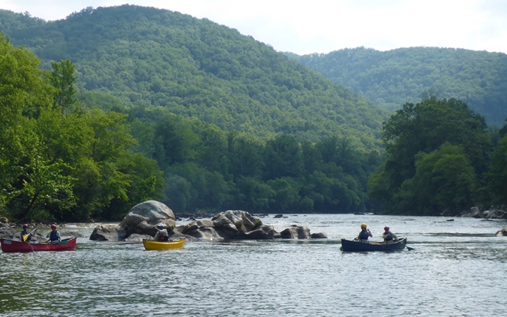 Three canoes, each carrying two people, float on a river amid rock formations. In the background, mountains are covered in thick green trees.